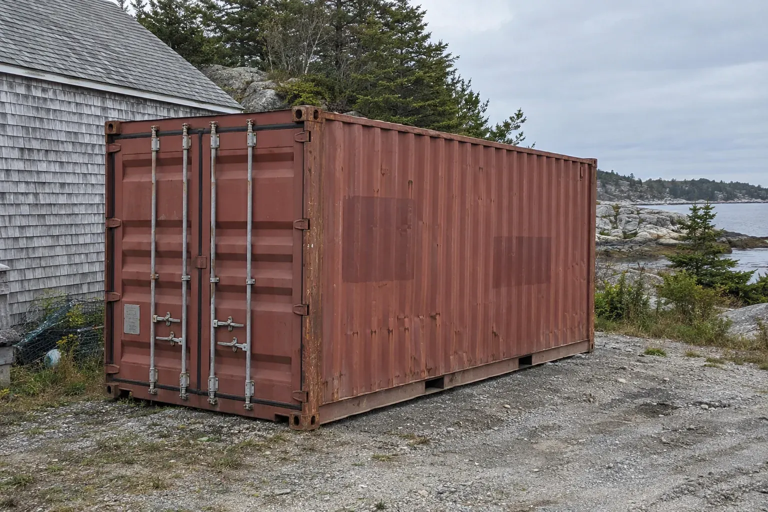 Used rust-red 20ft container beside a shingled shed near a rocky shore near Connecticut