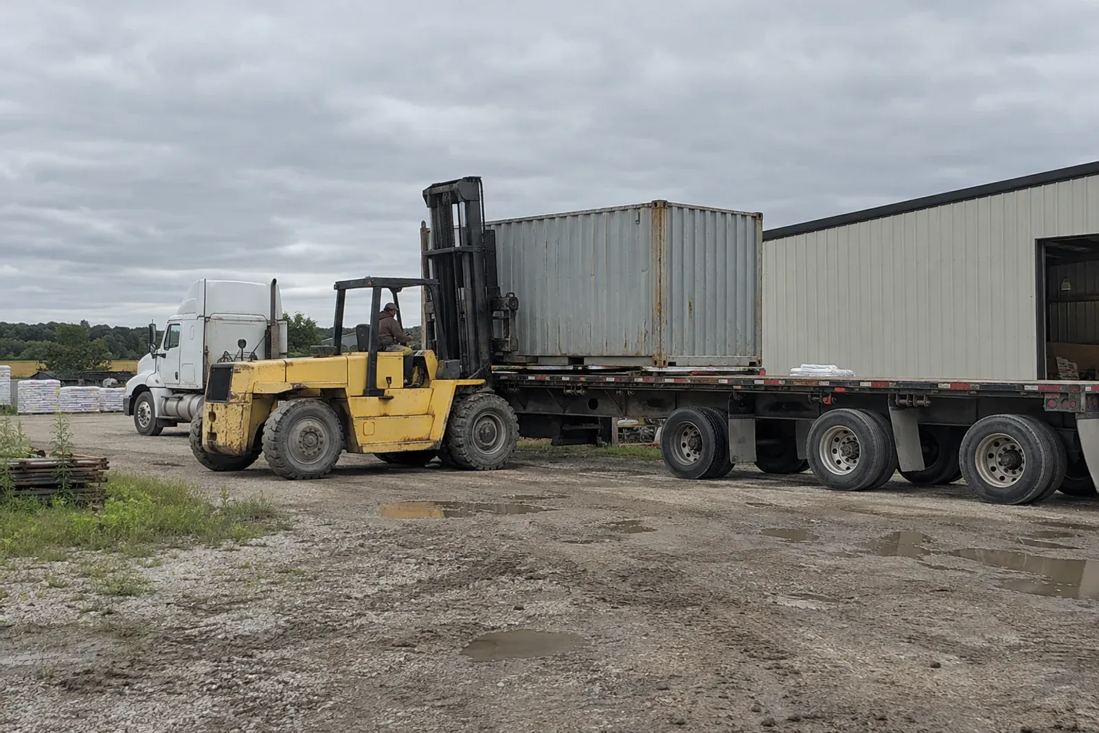 A forklift lifting a used 20 foot container off a flatbed semi trailer in a gravel yard